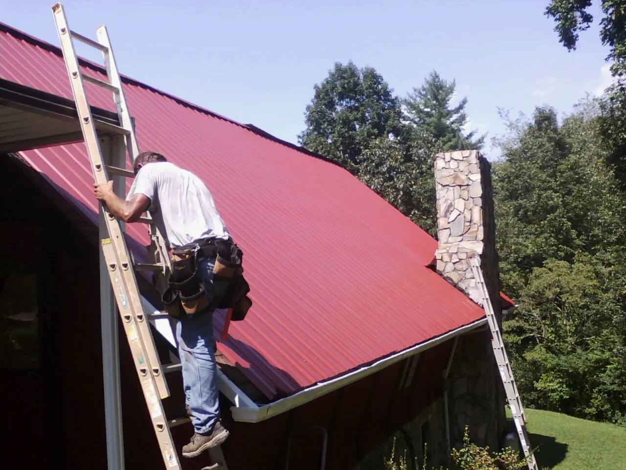 Roofer on a ladder beside a red metal roof and stone chimney — Soffit & Fascia Repair by Roofing Brothers in Pioneer